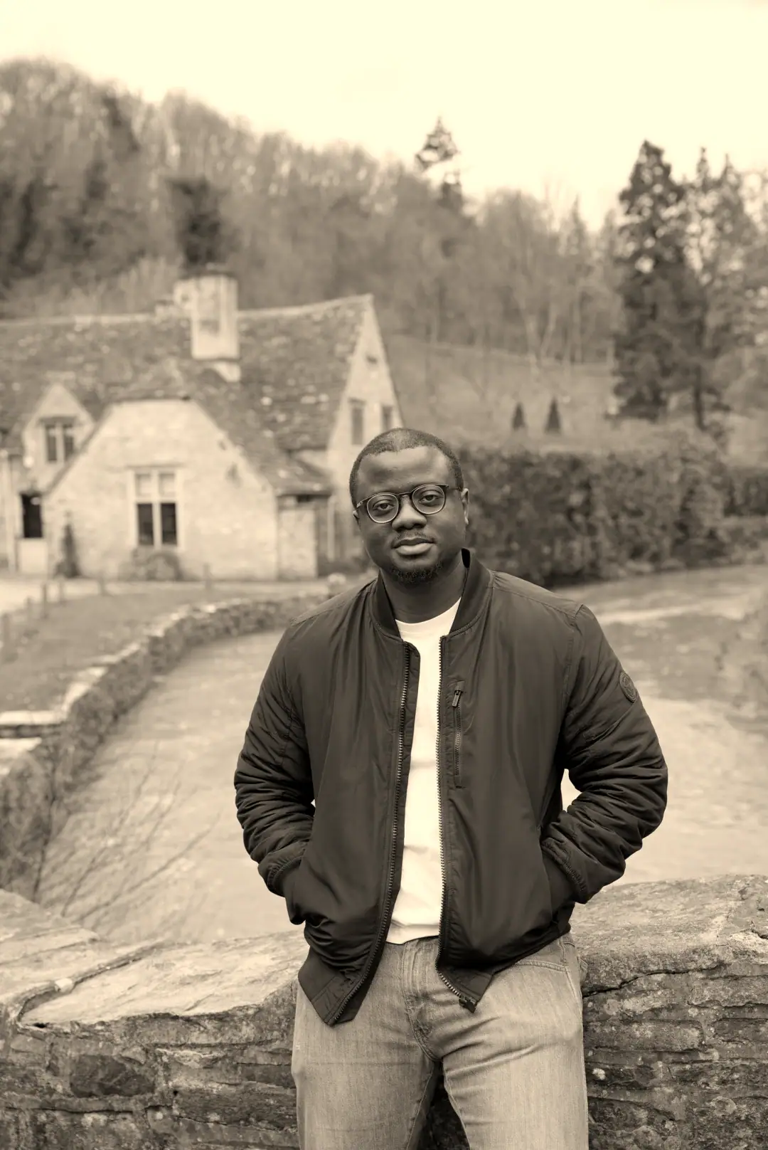 David leaning on a stone bridge in a Cotswolds village, a river and stone cottages behind him.