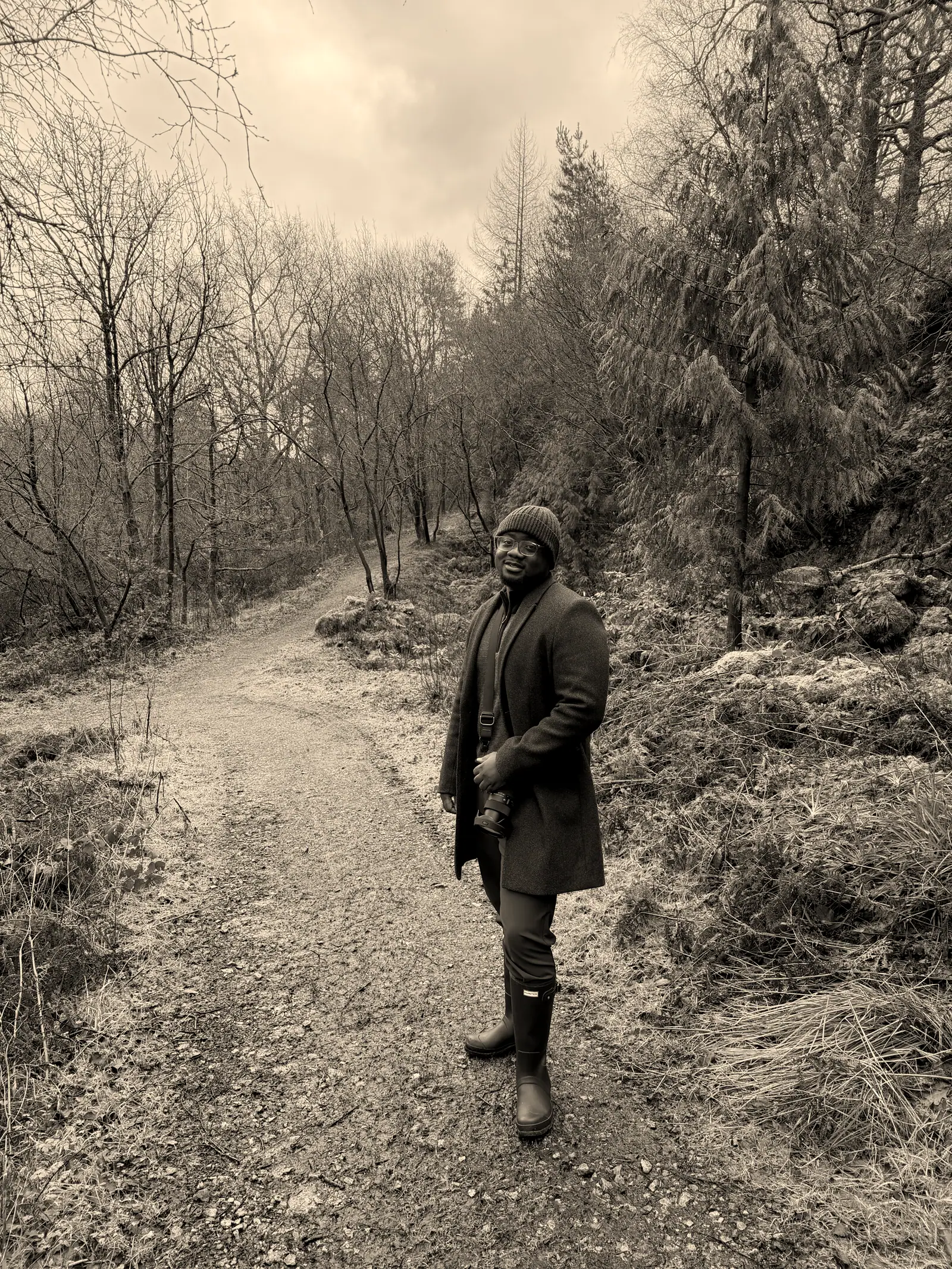 David on a woodland trail in a long coat and boots, camera in hand, looking back down the path.